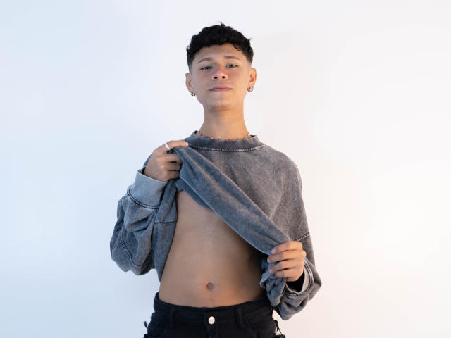 DanteEvans, shirtless, gray sweater lifted, exposing flat chest, small nipples, black pants, short black hair, light skin, small earrings, white background, standing, confident, slight smile.