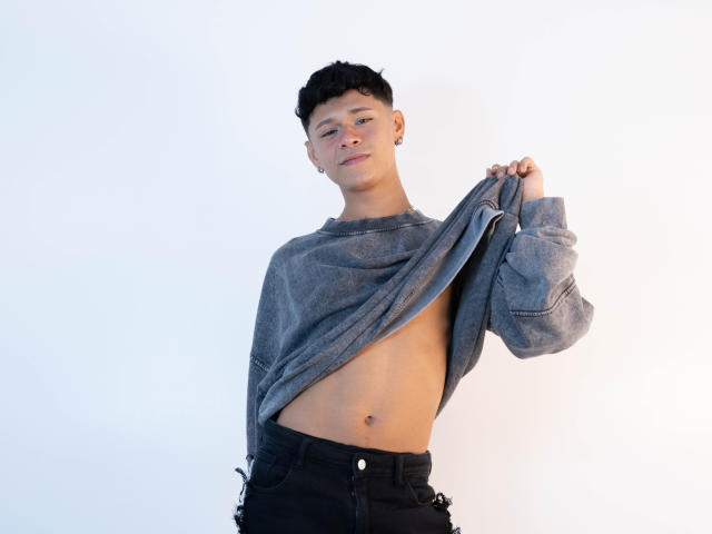 DanteEvans with short black hair, grey sweater lifted to show a flat, toned belly, wearing black pants, smiling slightly, small hoop earrings, white background.
