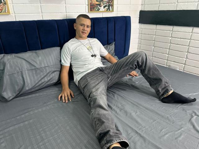 Lonny69 with shaved head, white t-shirt, grey jeans, black socks, and necklace, sitting on grey bed with blue headboard, smiling, hands on bed, legs spread.