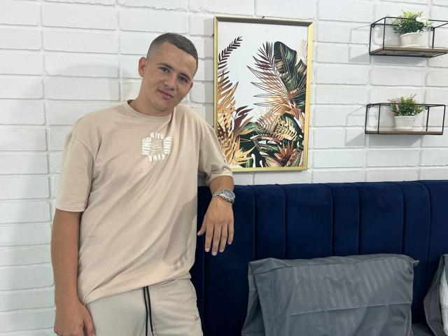 Lonny69 stands with a casual smile, wearing a beige t-shirt and matching pants, leaning on a blue velvet couch. He has short brown hair and a silver watch.