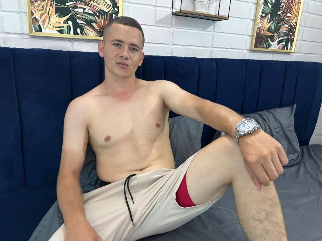 Shirtless Lonny69 with a slim build, short brown hair, and a watch, sits on a blue velvet bed, wearing beige shorts with a red bulge, looking at the camera.