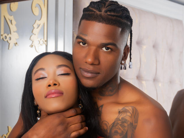 CataleyaAndEthan, un homme à la peau foncée avec des cheveux tressés et des tatouages, tient et embrasse une femme à la peau foncée, les yeux fermés et les lèvres brillantes. Tous deux sont torse nu, et la main de l'homme est posée sur son cou..