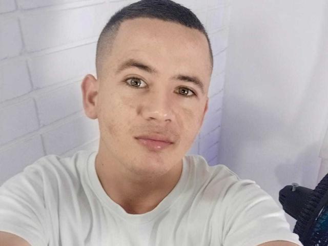 Lonny69 with a shaved head, green eyes, light skin, wearing a white tee, slight smile, short brown eyebrows, subtle acne on cheeks, standing against a white brick wall.