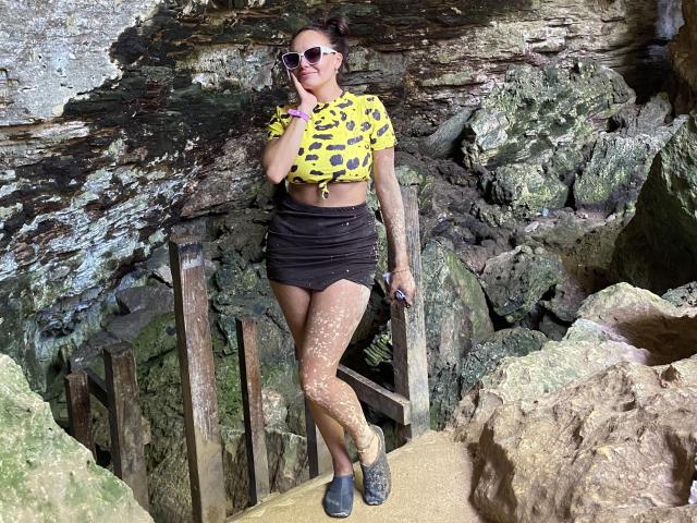 GalaMalkova with tan skin, tattooed arms, wearing a yellow polka-dot top, black mini-skirt, sunglasses, holding phone, standing on rocky path, legs covered in white sand, black shoes.