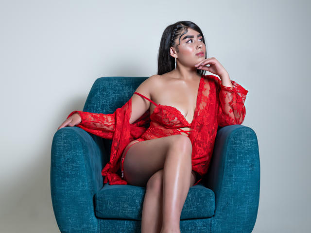 JulieAddams with medium tits, wearing red lace lingerie and sheer robe, sitting on teal chair, legs crossed, right hand on chin, looking to the side, dark hair, white wall.