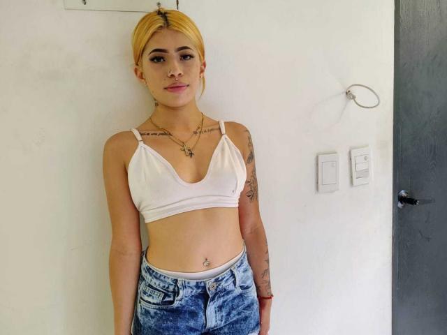 Emilly Hernandez, com cabelo loiro, blusa cropped branca, mostrando os seios, tatuagens nos braços, jeans azuis, piercing no umbigo, em pé em frente a uma parede branca..