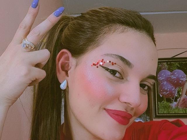 JulietaFoster69 has blue nails, a red face, glitter on her cheek, and a diamond ring, smiling with pink lipstick, brown hair in a ponytail, and a heart earring.