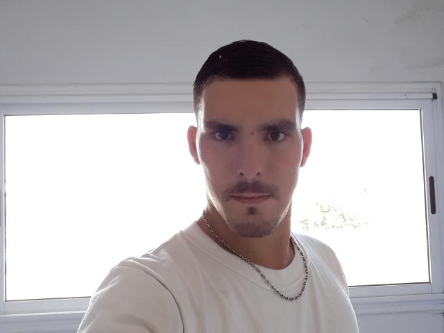ExedeNieve has a clean-shaven face, short dark hair, a mustache, and a slight goatee. He's wearing a white tee and a thin chain necklace, looking serious.