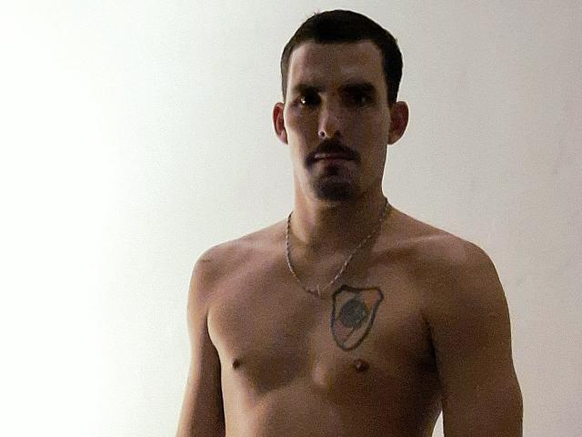 ExedeNieve is a shirtless dude with a tattoo on his left pectoral, wearing a chain necklace, looking serious with dark short hair and a small mustache.
