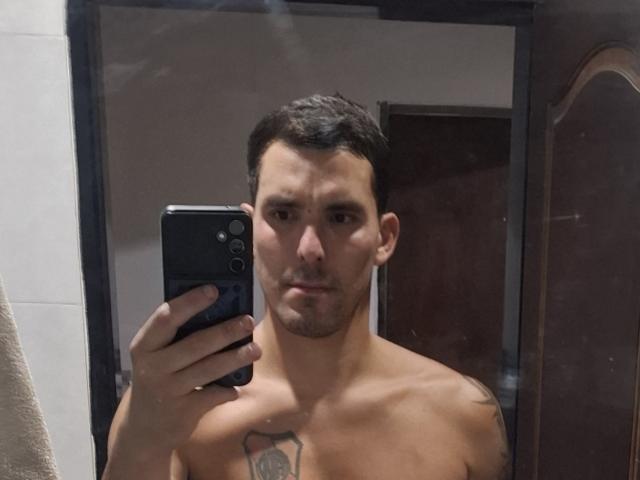 Shirtless ExedeNieve with short dark hair, light skin, and a chest tattoo, holding a black phone, taking a mirror selfie, showing a neutral expression.