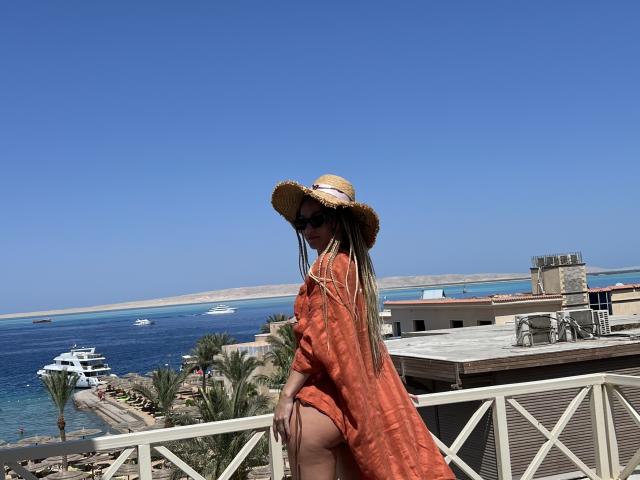 Lilitraya in an orange dress, straw hat, and sunglasses, showing her bare ass, standing on a rooftop with a clear blue sky and ocean in the background.