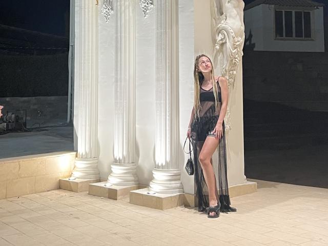 Lilitraya with long braids, black sheer dress, black high heels, holding black clutch, leaning against ornate white columns, smiling, showing her ass, at night.