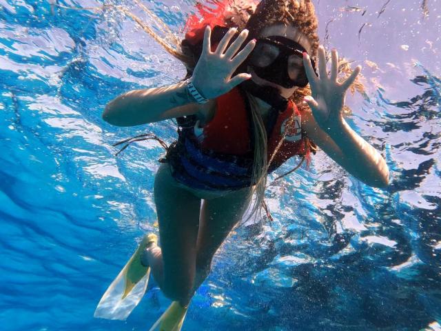 Lilitraya underwater, wearing a red life jacket, black mask, blue straps, yellow flippers, and a red shirt, with hands up, water rippling, and long brown hair flowing.