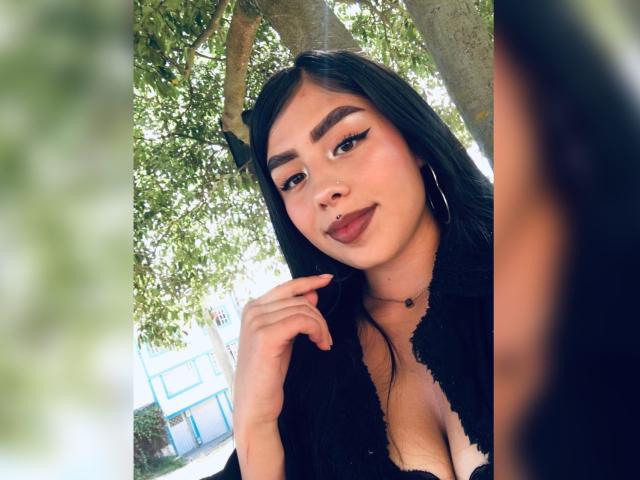 JulieAddams with long black hair, thick eyebrows, eyeliner, and red lipstick, wearing a black lace top, showing her cleavage, and a small nipple piercing, smiling.