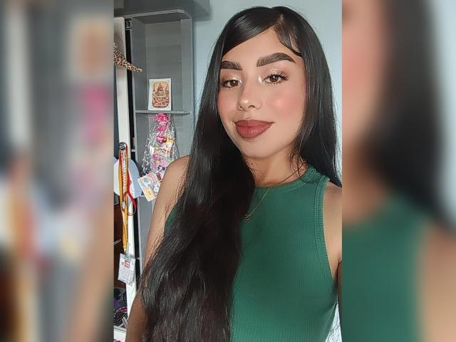 JulieAddams with long black hair, green sleeveless top, brown eyebrows, and dark red lipstick, standing in a mirror selfie, showing her tits slightly. Reflection on the right.
