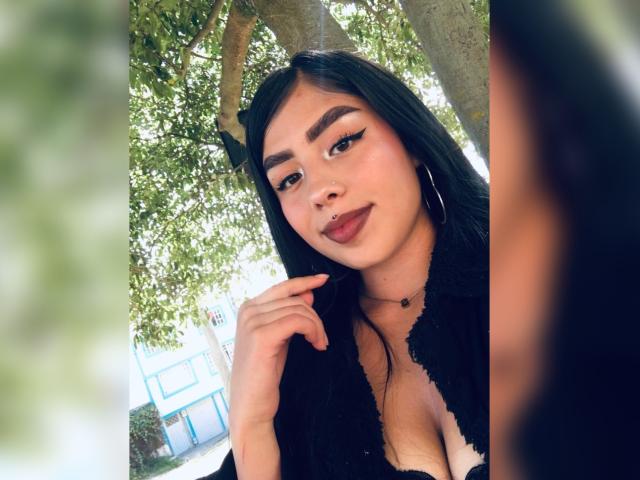 Julie Addams mit dunklem Haar, dicken Augenbrauen, Eyeliner im "Flügel"-Stil, einem schwarzen Spitzenoberteil und Reifenohrringen, lächelnd, mit dem Finger am Kinn, wobei ihr Brustpiercing zu sehen ist..