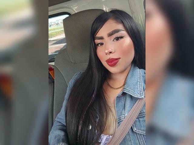 Julie Addams, con cabello negro largo, lápiz labial rojo y una chaqueta vaquera, sentada en un coche, con el cinturón de seguridad puesto, tiene un piercing en la nariz y una cadena de oro. Persona borrosa en primer plano..
