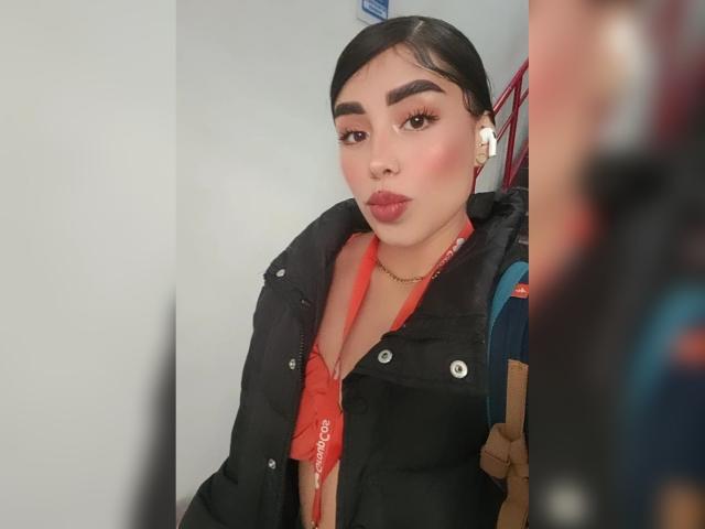 JulieAddams with dark hair, thick eyebrows, and red lipstick, pouting, wearing a black puffy jacket, red shirt, and earbuds, standing slightly angled with a blurred figure in front.