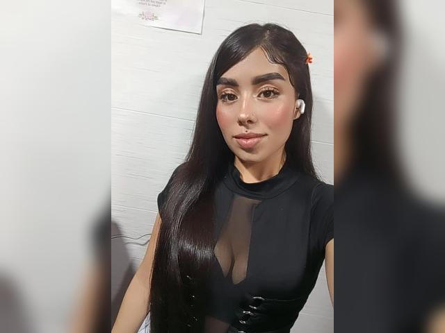 Julie Addams, avec de longs cheveux noirs, portant un haut noir transparent, laissant entrevoir de petites seins, avec des écouteurs blancs, souriant doucement, reflétée dans un miroir, avec une personne floue devant elle..