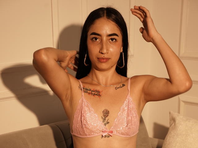 NoaMorena, con la pelle abbronzata, i capelli scuri e gli orecchini a cerchio, indossa un reggiseno di pizzo rosa, con le braccia alzate, mostrando tatuaggi sul petto e sotto il seno, in piedi sotto la luce calda del sole..
