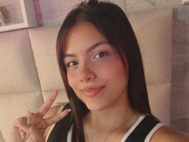 Emily Roberts tiene el pelo castaño, piel clara y está haciendo el gesto de la paz con su mano derecha. Lleva una blusa negra y blanca y está sonriendo ligeramente..