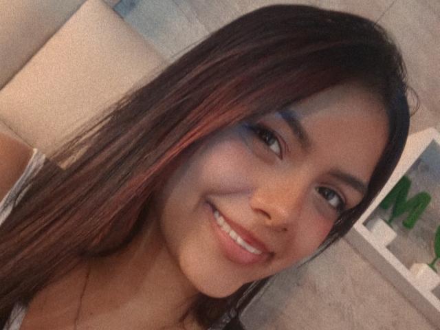 Emily Roberts tem um sorriso adorável, com olhos castanhos, cabelos longos castanhos com mechas vermelhas, e veste uma blusa cinza. Ela tem um nariz pequeno e está olhando para a câmera..