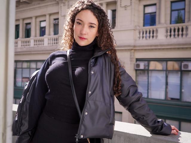 KristinaDubois : belle, avec des cheveux bruns bouclés, un pull noir à col roulé et une veste noire, appuyée contre un mur de béton, regardant l'appareil photo avec un léger sourire, la lumière du soleil sur son visage..