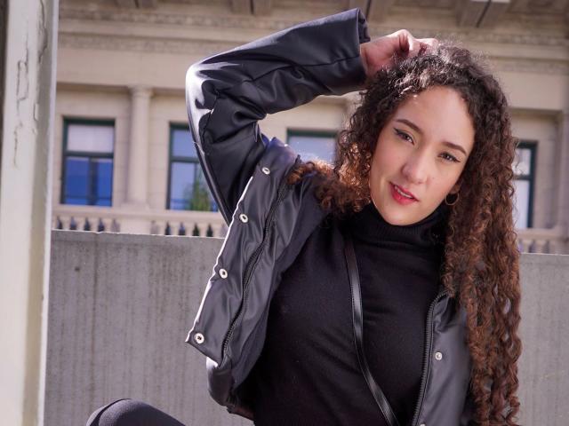 KristinaDubois - atraente, com cabelo castanho encaracolado, gola alta preta e jaqueta preta, com a mão no cabelo, sentada, sorrindo, mostrando seios pequenos, bolsa transversal preta, prédio branco ao fundo..