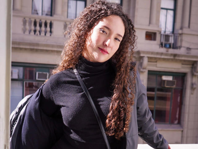 KristinaDubois - atraente, com cabelos castanhos cacheados, vestindo uma gola alta preta, jaqueta cinza e alça preta, sorrindo para a câmera, segurando uma mochila, dia ensolarado..