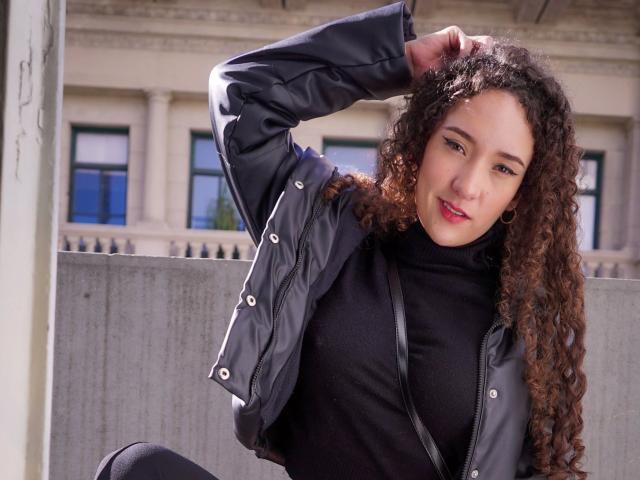 KristinaDubois-hot tem cabelos castanhos cacheados, veste uma gola alta preta e um casaco preto, com o braço levantado, mostrando seus pequenos seios, e um sorriso confiante e sexy..