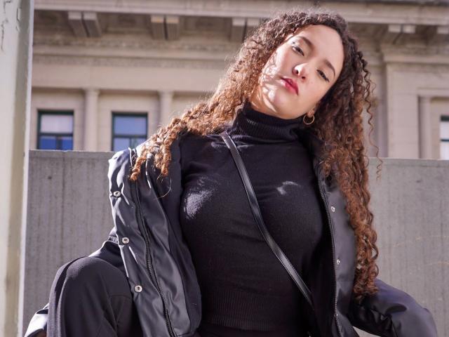 KristinaDubois – attraktiv mit lockigem, braunem Haar, schwarzem Rollkragenpullover und schwarzer Jacke, blickt nach unten, rote Lippen, mittelgroße Brüste, schwarze Umhängetasche, goldene Ohrringe im Stecker-Design, steht im Sonnenlicht..