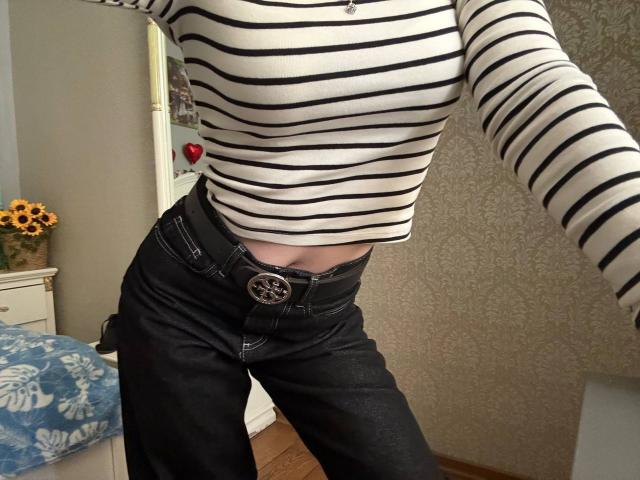 Siyana's striped long-sleeve shirt, black jeans with a silver belt, and visible tan midriff. Her jeans are slightly unbuttoned. Sunflowers on a white dresser.