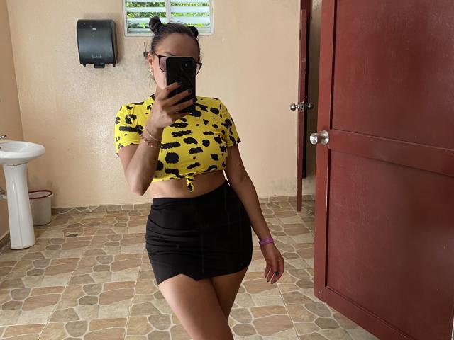 GalaMalkova usando uma cropped amarela, estampa de leopardo preta, saia mini preta, segurando um celular, usando óculos, pulseira rosa, em pé em um quarto com piso de azulejos, porta à direita, pia à esquerda..