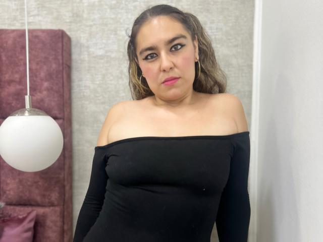 TaylorNova mit welligem, braunem Haar, trägt ein schwarzes Oberteil mit einem Schulterauschnitt, hat kleine Brüste, pinken Lippenstift und einen neutralen Gesichtsausdruck und steht vor einer weißen Wand..