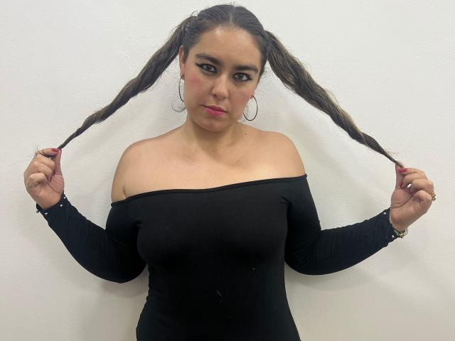 TaylorNova, avec ses cheveux bruns bouclés attachés en deux couettes, porte une robe noire à une épaule, se tient les cheveux, dévoilant sa poitrine, avec du rouge à lèvres, des boucles d'oreilles cerceaux, et un fond de mur blanc..