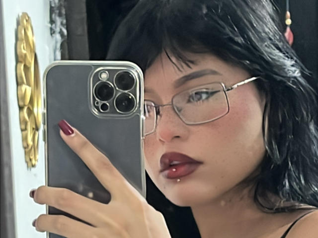 ElizabethRoux with black hair, glasses, and red lipstick, holding a smartphone, showing her red-manicured fingers and pierced lower lip, looking at the camera.