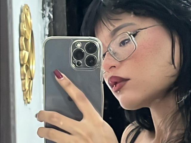 Elizabeth Roux, con gli occhiali, i capelli neri e un piercing al labbro, sta scattando un selfie con un iPhone grigio, mostrando le unghie rosse e la bocca leggermente aperta. Le sue natiche non sono visibili..