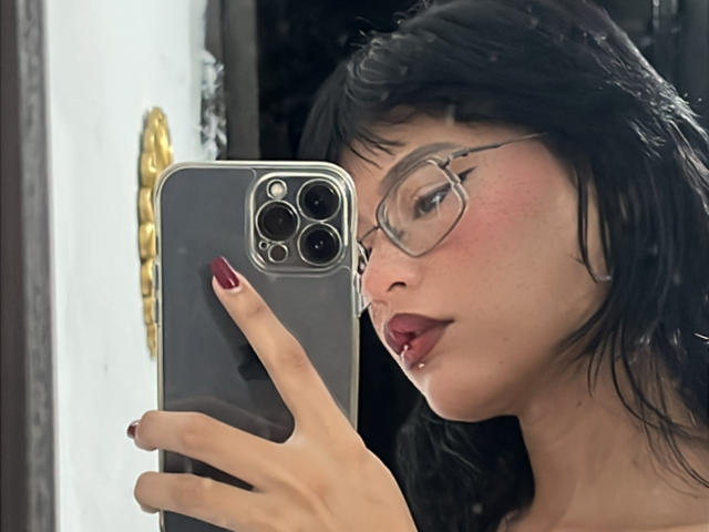ElizabethRoux with black hair, glasses, and red lipstick, holding a gray iPhone with red nails, taking a selfie, showing a slight pout and lip piercing.