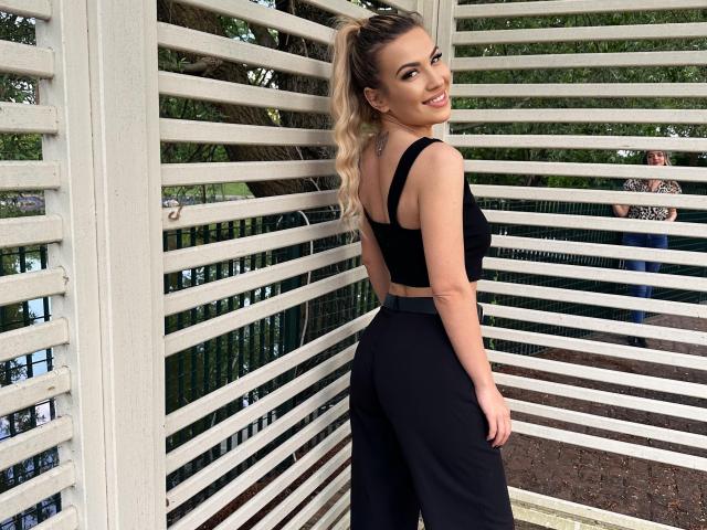 ZoeyMarie with blonde ponytail, black crop top, and black high-waisted pants, showing her ass, smiling, standing in front of white slatted wall, green trees in background.