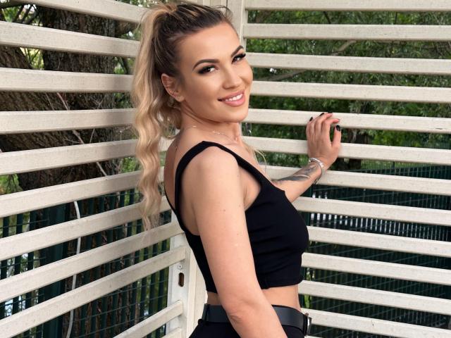 ZoeyMarie with blonde ponytail, smiling, wearing black crop top, showing her tits, standing against white slats, right hand on slats, tattoo on left arm, outdoor setting.
