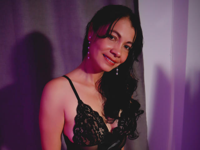 MelissaBaud with dark wavy hair, wearing black lace lingerie, smiling, earrings, pinkish-purple light, shadow on wall, visible tits, cute, attractive, soft lighting, relaxed pose.