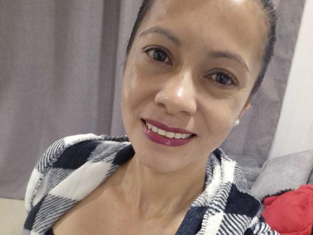 MelissaBaud has tan skin, brown eyes, and dark hair in a bun. She's smiling with red lipstick, wearing a checkered top, and has small, round earrings.