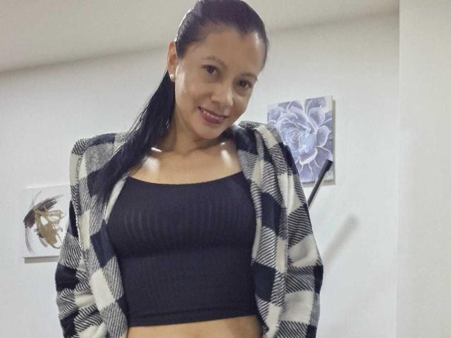 MelissaBaud with long black hair, wearing a black ribbed crop top and a black-and-white checkered jacket, smiling at the camera, showing her small tits and tan skin.