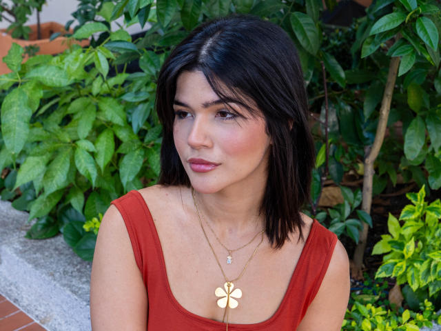 ScarlletZhen hat dunkelbraunes Haar, ein rotes Kleid, dezentes Make-up, rosafarbene Lippen, eine goldene Halskette mit einem Blumenanhänger und einen grünen, blattigen Hintergrund. Ihr Gesäß ist nicht sichtbar..