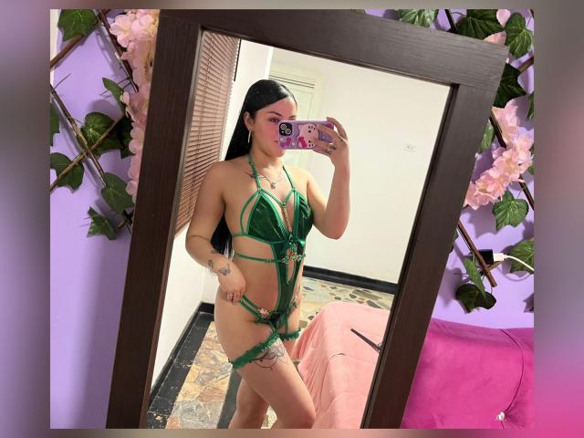 EmmieX with long black hair, green strappy lingerie, showing small tits, a tattoo on her thigh, taking a mirror selfie, holding a pink phone with floral case.