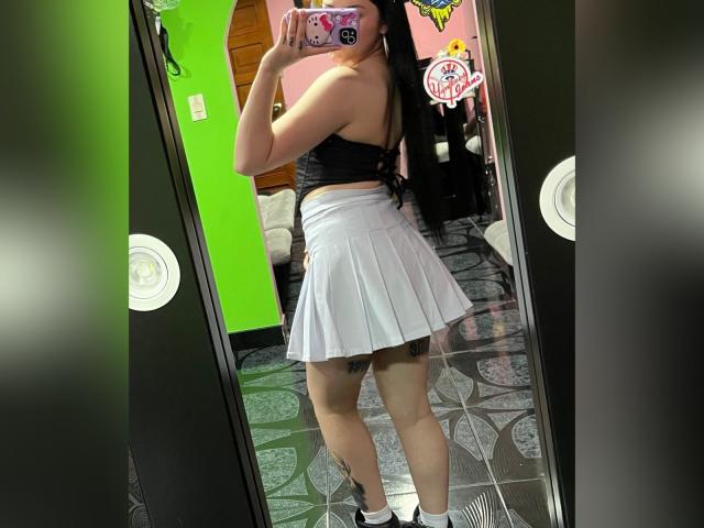EmmieX with long black hair, wearing a black strapless top and white pleated skirt, showing her ass in a mirror selfie. She has tattoos on her thigh.