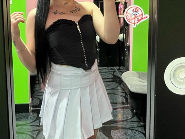 EmmieX with black strapless corset, white pleated skirt, long black hair, tattoo on chest, holding phone in mirror selfie. Visible boobs, no ass. Green wall in background.