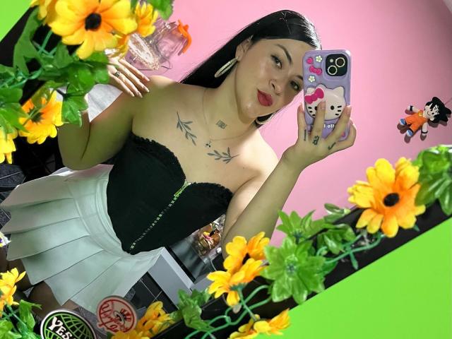EmmieX with long black hair, red lipstick, black strapless corset, white skirt, and tattoo on chest, taking a mirror selfie with sunflowers, holding a phone with a cute case.