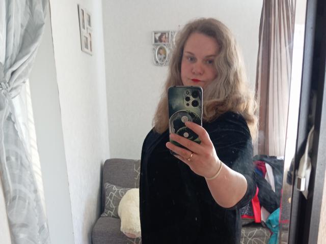 Mavka69 with wavy blonde hair, wearing a black top, taking a mirror selfie with a smartphone, showing her reflection, medium tits, and a slightly annoyed expression.