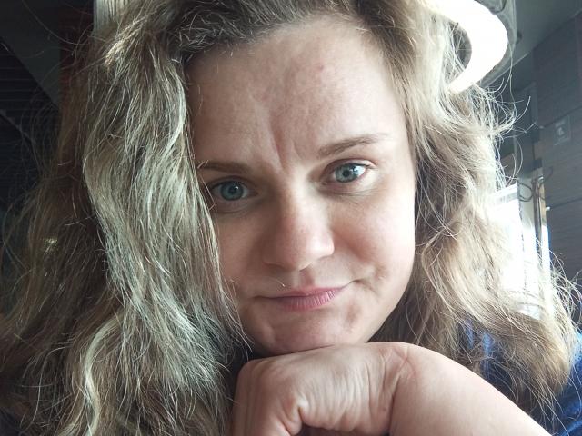 Mavka69 a les cheveux blonds ondulés, les yeux bleus et un sourire discret. Elle repose son menton sur sa main, et on peut voir un anneau dans son nez ainsi qu'une petite imperfection sur son front..